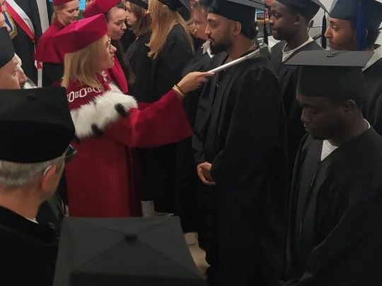 Ceremonia akademicka w nowoczesnej sali. Osoby w czarnych togach i biretach stoją w rzędzie, a kobieta w czerwonej todze z gronostajem pasuje ich dotykając ramienia insygnium. W tle widoczny ekran i pozostali goście