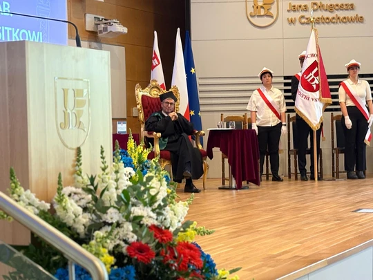 Na zdjęciu widzimy uroczystość w Uniwersytecie Jana Długosza w Częstochowie. Centralnie, na podwyższeniu, siedzi osoba w stroju akademickim na ozdobnym fotelu. Obok stoi stół nakryty bordowym obrusem oraz kilka flag W tle stoją młodzi ludzie ze sztandarem.