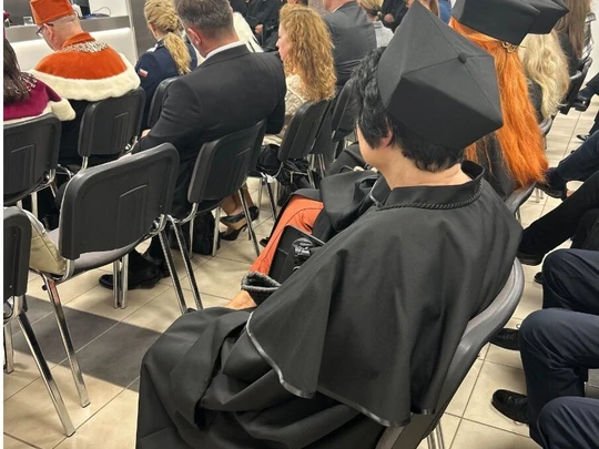 Nowoczesna sala podczas uroczystości akademickiej. Przed widownią osoba wygłasza przemówienie. Wśród gości widoczni absolwenci w czarnych togach i biretach. Atmosfera oficjalna i skupiona.