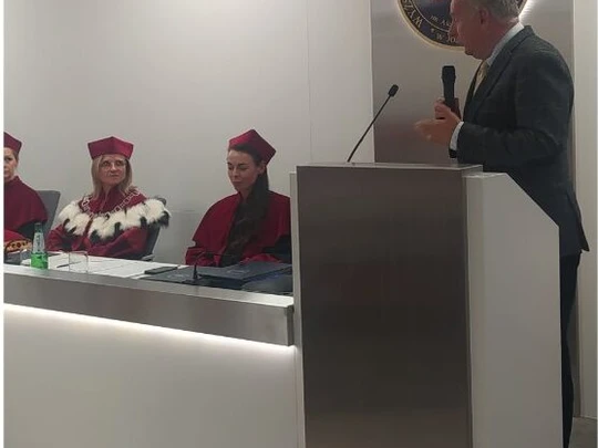Sala akademicka podczas inauguracji roku akademickiego. Mężczyzna w garniturze przemawia przy mównicy. Po lewej stronie za stołem siedzą dwie osoby w czerwonych togach i biretach. W tle srebrny emblemat uczelni z biało-czerwoną flagą. Atmosfera formalna.