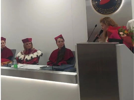 Kobieta z bukietem kwiatów stoi przy mównicy i przemawia podczas akademickiej uroczystości. Po lewej komisja w czerwonych togach i biretach za stołem. W tle emblemat uczelni z biało-czerwoną flagą.