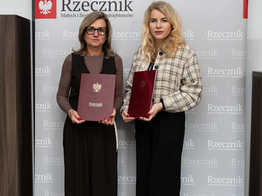 Na jasnym tle, przed ścianą z napisem „Rzecznik” i czerwonym symbolem orła, stoją dwie osoby, trzymające bordowe teczki lub dyplomy z polskim godłem. Ujęcie oficjalne, instytucjonalne.