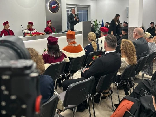 Nowoczesna sala konferencyjna wypełniona gośćmi i pracownikami akademickimi w ceremonialnych togach. Przy mównicy stoi przemawiający, przed stołem prezydialnym siedzą osoby w kolorowych togach. Na ścianie emblemat uczelni.