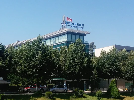 Nowoczesny budynek uniwersytetu otoczony drzewami. Na dachu widoczny napis „UNIWERSYTET” oraz flaga. Jasne, słoneczne niebo i zielona roślinność w pierwszym planie.