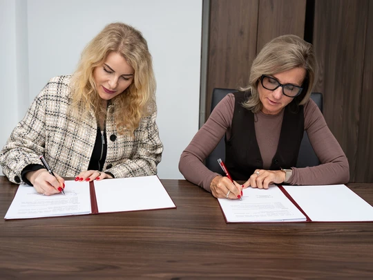 Dwie kobiety siedzą przy stole i podpisują dokumenty. Na stole leżą białe arkusze papieru oraz czerwone długopisy. Tło stanowią jasna ściana i drewniane panele. Atmosfera jest oficjalna i formalna.