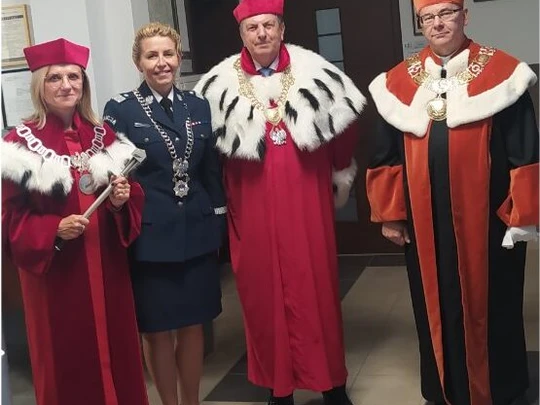 Cztery osoby pozują podczas oficjalnej uroczystości w budynku uczelni. Dwie postacie w czerwonych togach z wykończeniem gronostajowym i beretach, jedna w pomarańczowo-czarnej todze ceremonialnej, czwarta w granatowym mundurze z łańcuchem rektorskim.