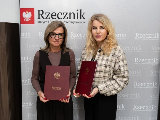 Na jasnym tle, przed ścianą z napisem „Rzecznik” oraz czerwonym godłem Polski, stoją dwie kobiety, trzymające bordowe teczki lub dyplomy z wytłoczonym orłem. Ujęcie oficjalne, instytucjonalne.