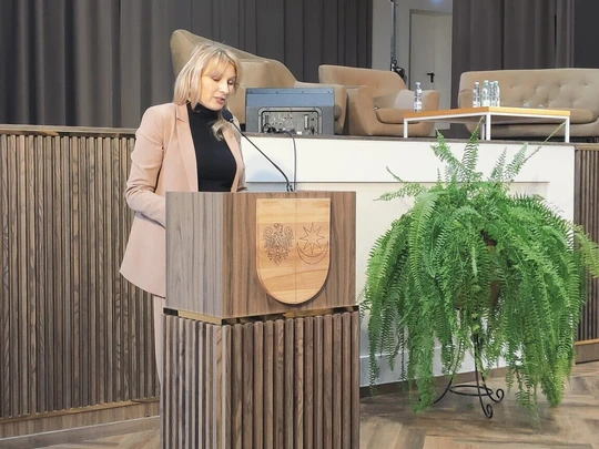 Kobieta stoi przy mównicy w jasnej sali konferencyjnej. W tle wysokie zasłony, podium z rośliną i fotele. Nad sceną godło Polski. Oświetlenie sceniczne, atmosfera oficjalna i profesjonalna.