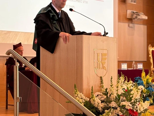 Na zdjęciu widać osobę ubraną w togę i biret akademicki, stojącą za mównicą i przemawiającą podczas uroczystości w dużej, jasnej sali. Przed mównicą widoczne są kwiaty, a w tle siedzą inne osoby również ubrane w togi. Całość wskazuje na oficjalny charakter