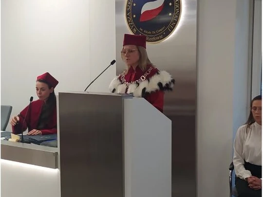 Sala akademicka z osobą w czerwonej todze i birecie wygłaszającą mowę przy mównicy, z tyłu widoczny srebrny emblemat z biało-czerwoną flagą. Po lewej członek komisji w todze, po prawej siedzi kobieta. Atmosfera oficjalna.