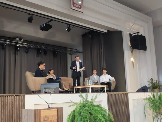 Na scenie z szarymi zasłonami cztery osoby siedzą na kanapach, jedna stoi z mikrofonem. W tle godło Polski. Oświetlenie sceniczne. Przód sceny drewniany, po prawej rośliny i ekran. Oficjalne wydarzenie, sala konferencyjna.