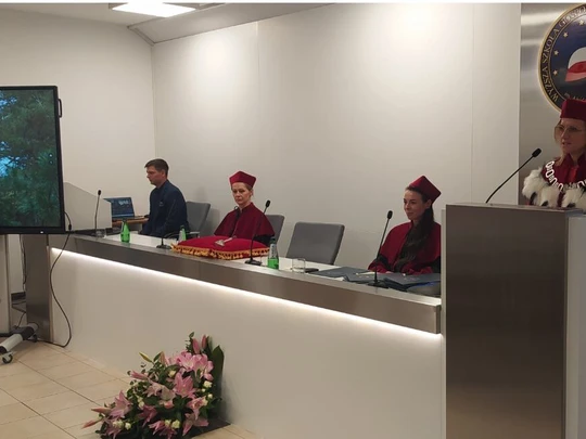 W nowoczesnej, jasnej sali akademickiej komisja w czerwonych togach i biretach siedzi za długim stołem. Po lewej stronie widoczny jest ekran z prezentacją na którym widać imię prowadzącej. Po prawej stronie mównica i uczelniany emblemat. Atmosfera formalna