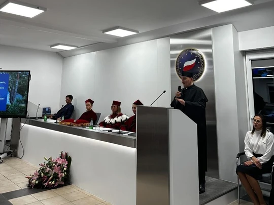 Nowoczesna sala, osoba w czarnej todze przemawia przy mównicy z mikrofonem. W tle emblemat uczelni z biało-czerwonym motywem. Po lewej komisja w czerwonych togach za stołem, z przodu bukiet kwiatów, po prawej elegancka kobieta.