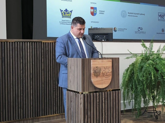 Mężczyzna w garniturze stoi przy mównicy na tle dużego ekranu z logotypami i napisem „Forum Edukacji Prawniczej”. Obok podium zielona roślina. Sala jasna, atmosfera oficjalna i profesjonalna.