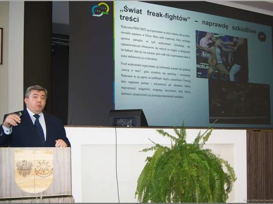 Mężczyzna w garniturze występuje przy mównicy w jasnej sali konferencyjnej. W tle wyświetlany jest slajd z tekstem i zdjęciami dotyczący „freak-fightów” w internecie, po prawej ekran z wizualizacją. Obok mównicy stoi zielona roślina.