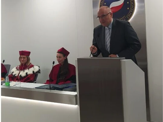 Sala akademicka podczas wystąpienia. Mężczyzna w garniturze przemawia przy mównicy. Po lewej stronie za stołem siedzą dwie osoby w czerwonych togach i biretach. W tle srebrny emblemat uczelni z biało-czerwoną flagą. Atmosfera formalna.