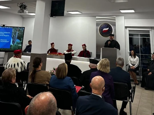 Uroczystość akademicka w nowoczesnej sali konferencyjnej. Na pierwszym planie siedzą słuchacze w eleganckich, ciemnych strojach, zwróceni w stronę podwyższenia. Przed nimi znajduje się podium, przy którym przemawia osoba w czarnym stroju akademickim