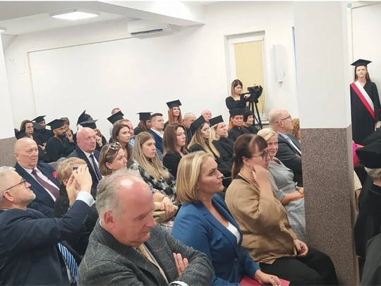 Widok sali podczas inauguracji roku akademickiego. W pierwszych rzędach siedzą absolwenci i goście, część w togach i biretach. Z prawej strony stoją trzy osoby ze sztandarem uczelni. Atmosfera oficjalna i podniosła.