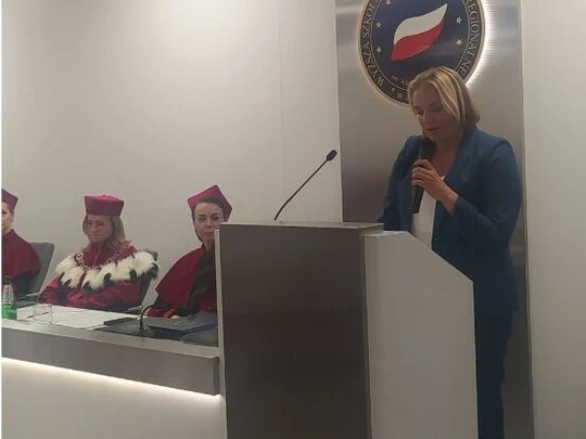 Kobieta stoi przy mównicy i przemawia podczas uroczystości akademickiej; po lewej trzy osoby w czerwonych togach i biretach. W tle emblemat z biało-czerwoną flagą. Sala jasna, atmosfera oficjalna i podniosła.