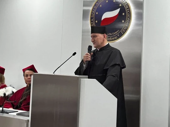 Osoba w czarnej todze przemawia przy mównicy w nowoczesnej auli. Na ścianie emblemat uczelni z polską flagą. Po lewej stronie przy stole siedzą osoby w czerwonych togach i biretach, atmosfera formalna i podniosła.