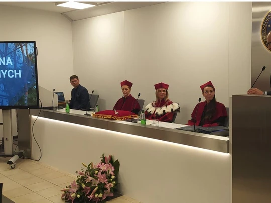 Sala akademicka, z lewej strony ekran z napisem „Wystąpienia zaproszonych gości”. Przy długim stole siedzą trzy osoby w czerwonych togach i biretach oraz jedna osoba w garniturze. Przy mównicy stoi mężczyzna. Atmosfera oficjalna.