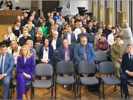 Widok na dużą salę konferencyjną wypełnioną uczestnikami. Kilkadziesiąt osób różnych płci siedzi w równych rzędach, przodem do fotografa. W tle jasne wnętrze, są złote balustrady, drewniana podłoga, okna po prawej stronie. Atmosfera oficjalna.