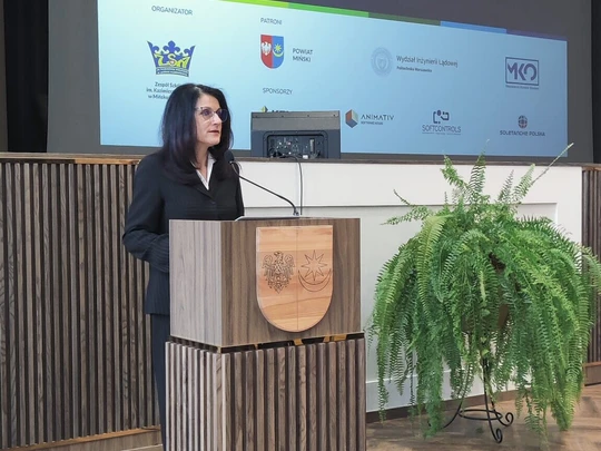 Kobieta w garniturze stoi przy mównicy na tle dużego ekranu z logotypami i napisem „Forum Edukacji Prawniczej”. Obok podium zielona roślina. Sala jasna, atmosfera oficjalna i profesjonalna.
